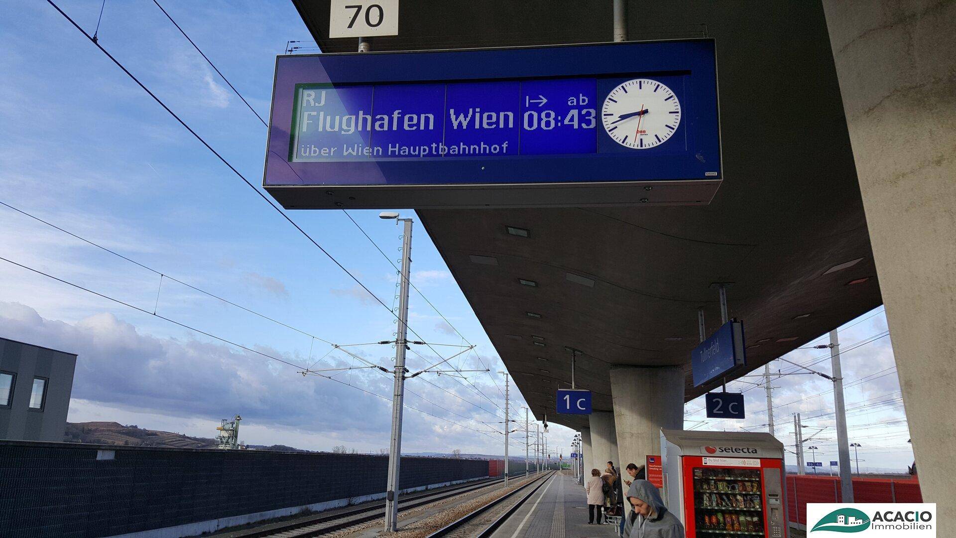Anzeigetafel am Bahnhof zeigt Abfahrtszeiten zum Flughafen Wien unter bewölktem Himmel.