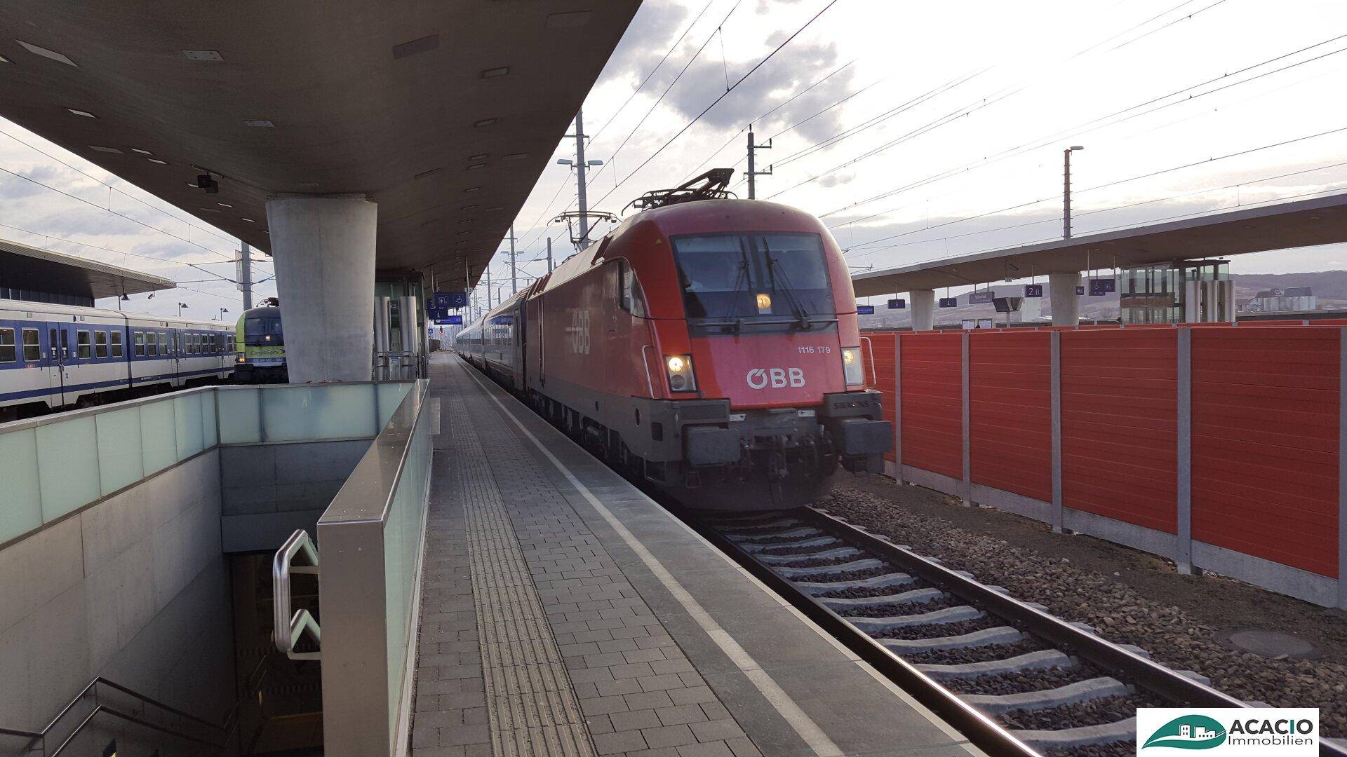 Roter Zug fährt in den modernen Bahnhof ein, mit Bahnsteigen und Oberleitung unter weitem Himmel.