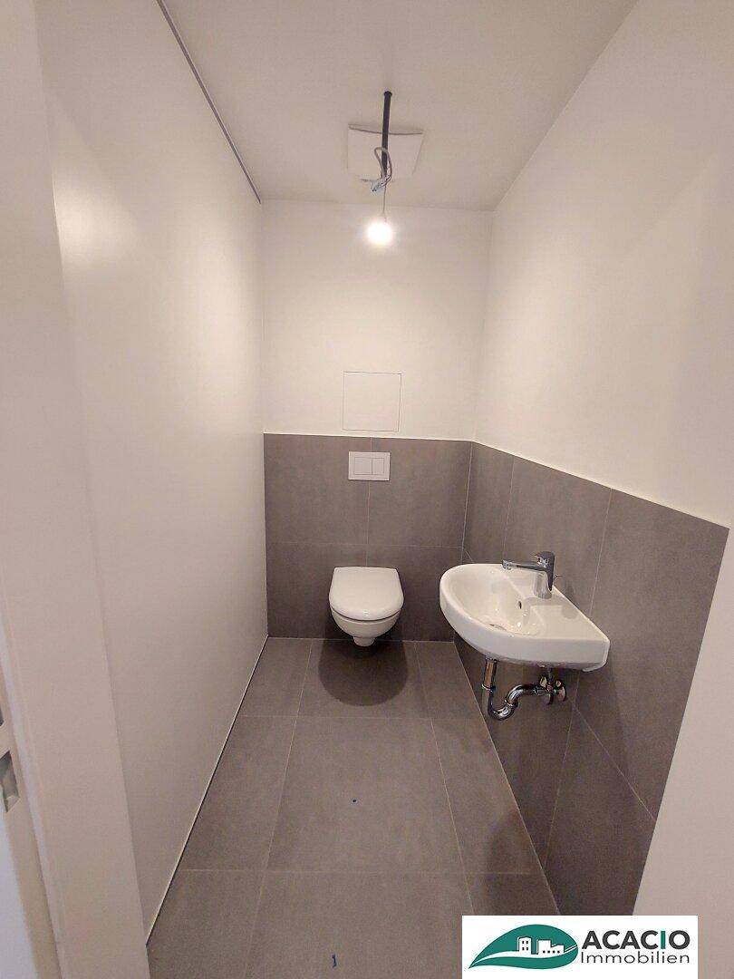 Modernes Gäste-WC mit wandhängender Toilette, kleinem Waschbecken und grauen Wandfliesen.