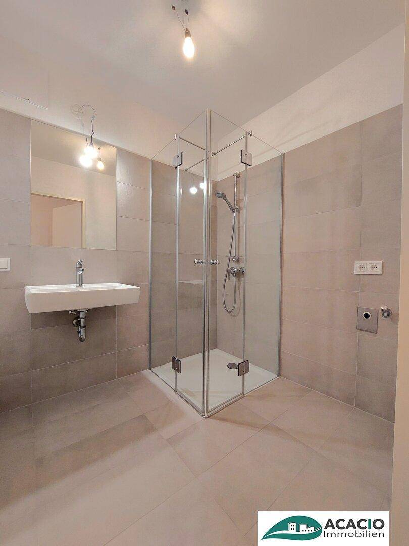 Modernes Badezimmer mit grauen Fliesen, wandhängendem Waschbecken und einer geräumigen Glasdusche.