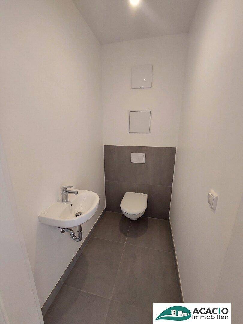 Gäste-WC mit wandhängender Toilette, kleinem Waschbecken und grauen Bodenfliesen.