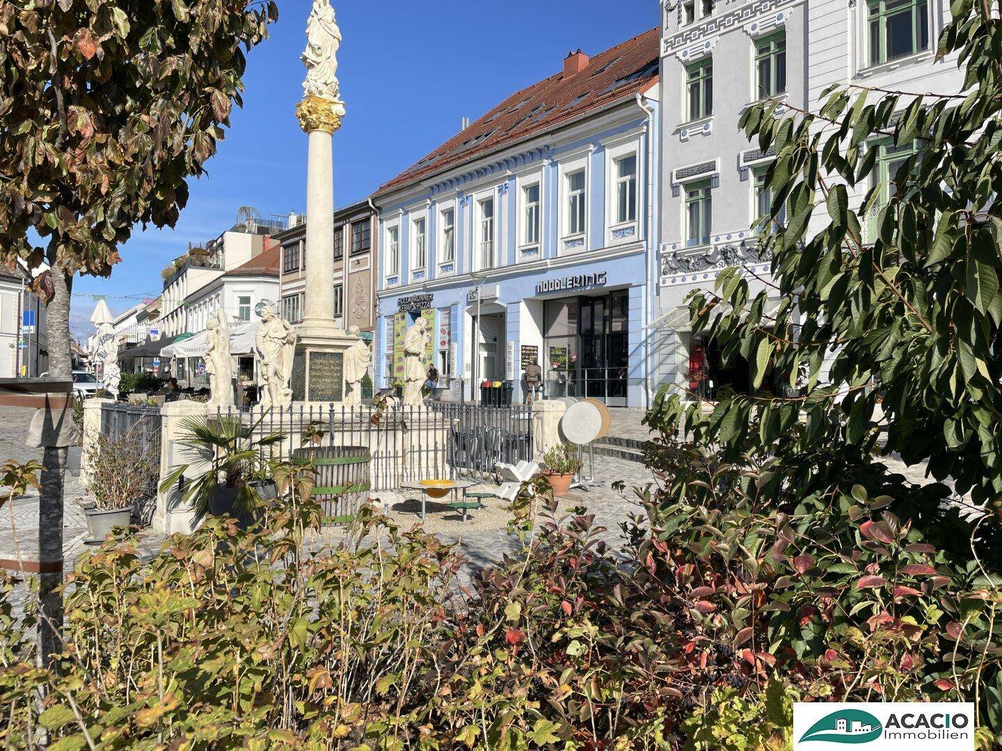 Historische Gebäude und ein Denkmal auf einem belebten Stadtplatz unter blauem Himmel.