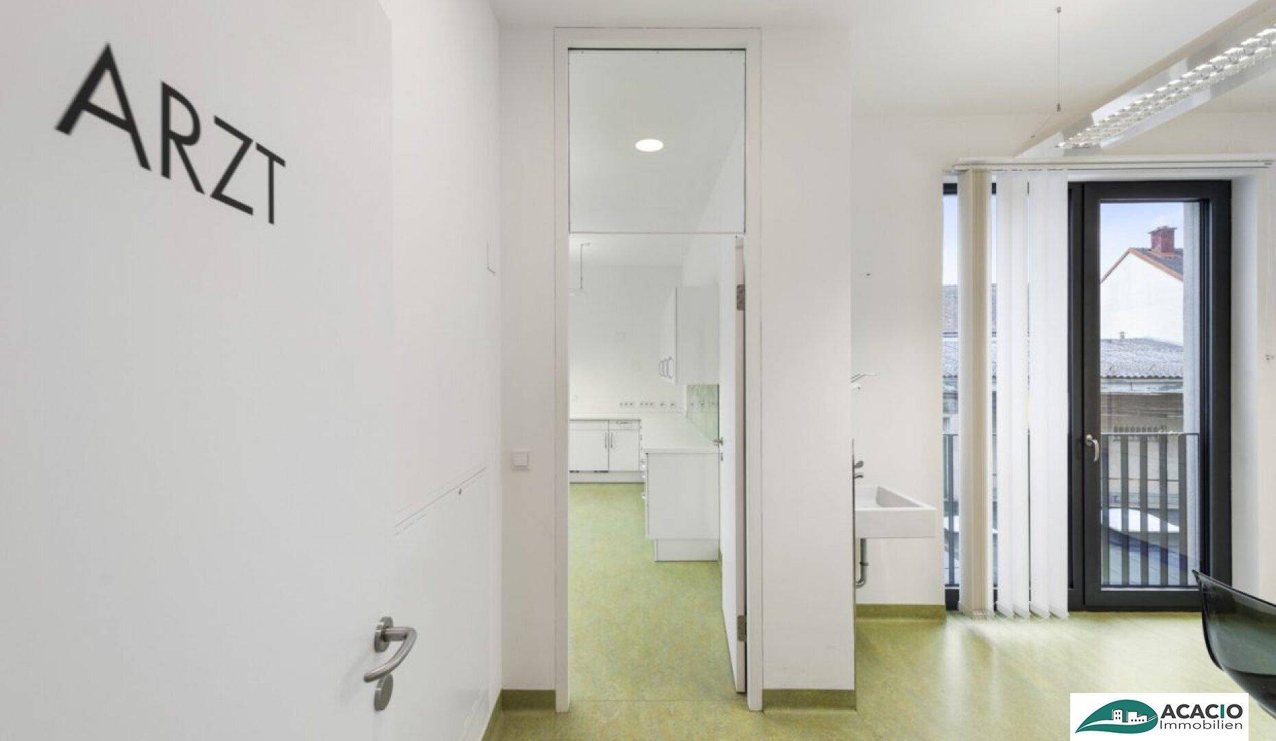 Blick in ein Behandlungszimmer einer Arztpraxis mit grünem Boden, weißer Einrichtung und Zugang zu einem Balkon.