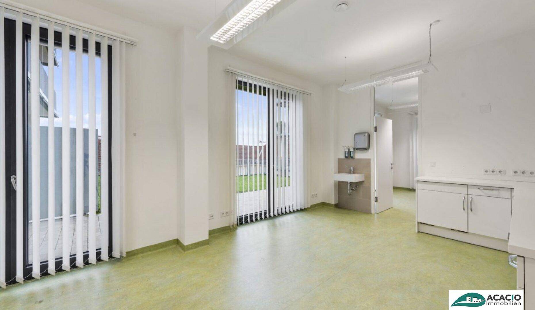 Geräumiger Behandlungsraum mit großen Fenstern, vertikalen Jalousien, grünem Boden und einem Waschbeckenbereich.