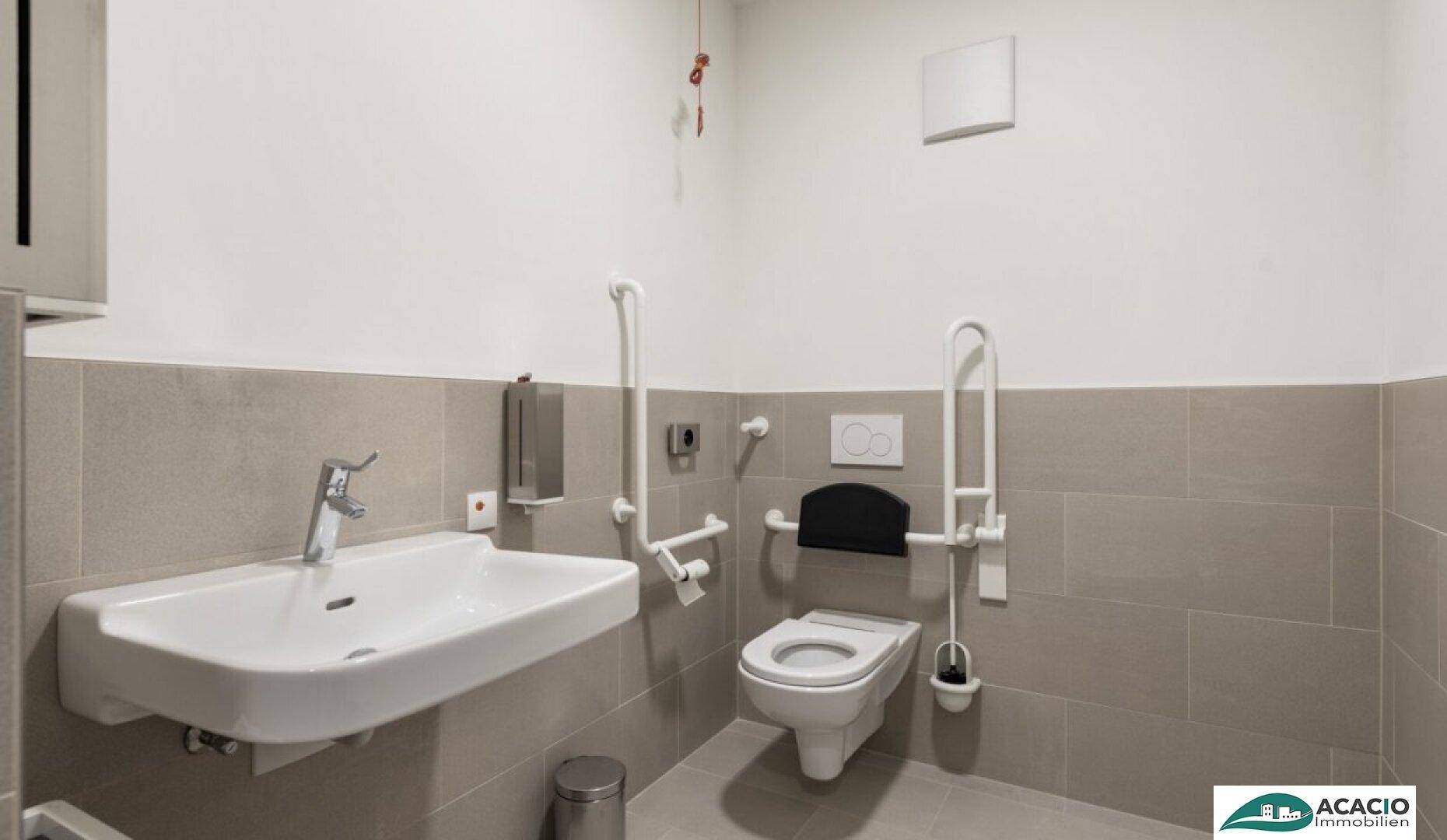 Barrierefreies Badezimmer mit Haltegriffen, wandhängendem WC und Waschbecken, sowie hellgrauen Fliesen.