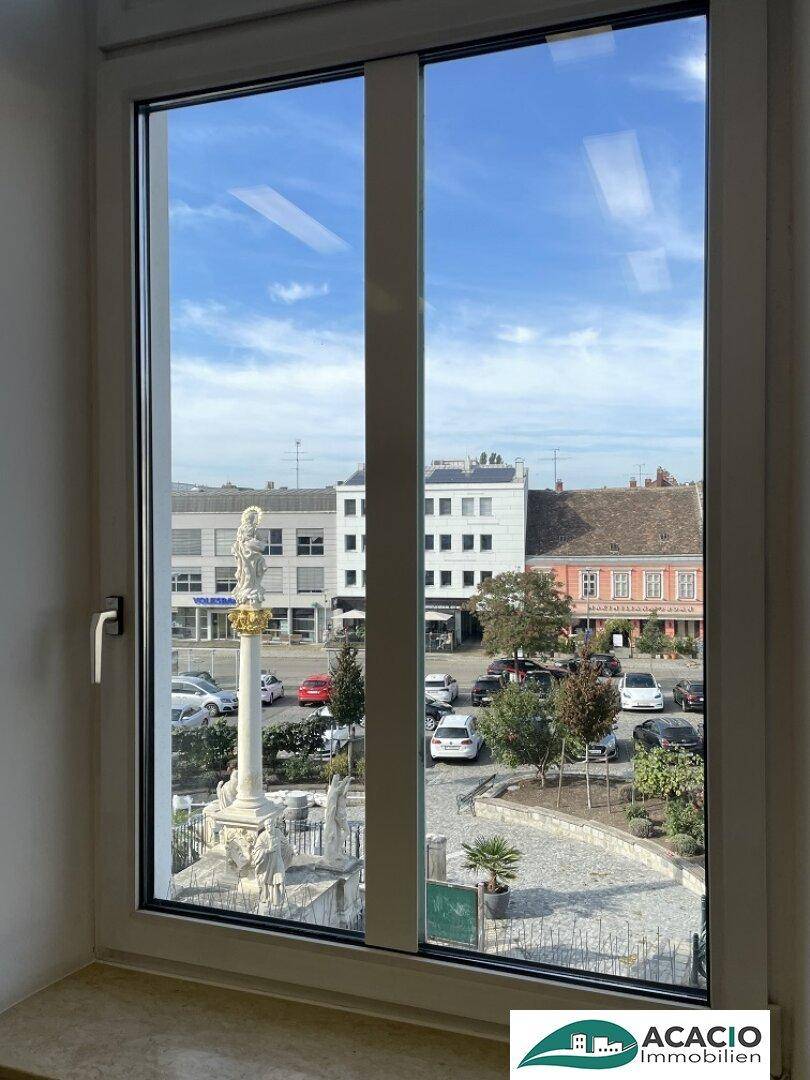 Fensterblick auf einen historischen Stadtplatz mit Denkmal und umliegenden Gebäuden.