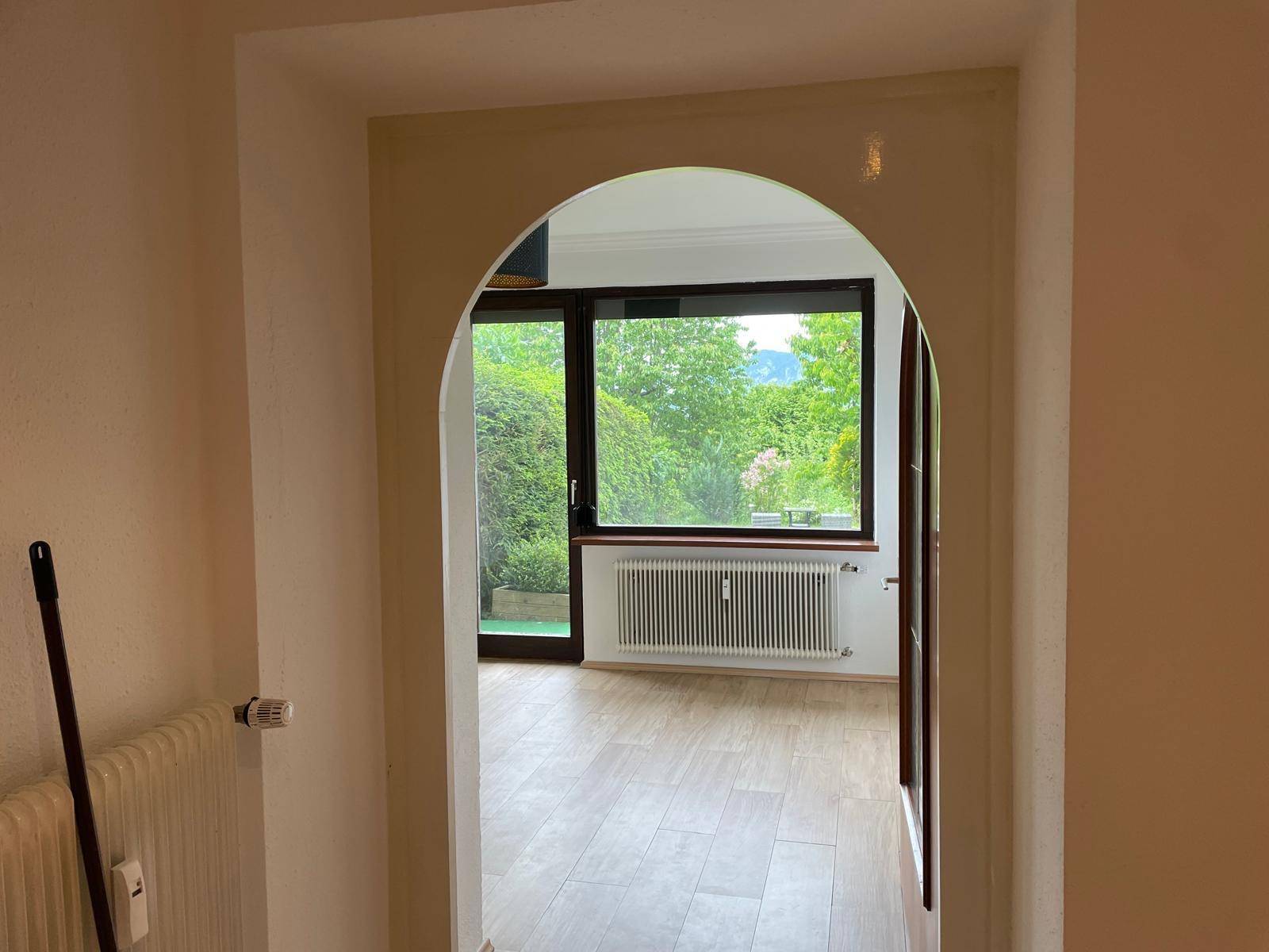 Blick durch einen Rundbogen in einen hellen Raum mit Holzboden und grüner Aussicht.