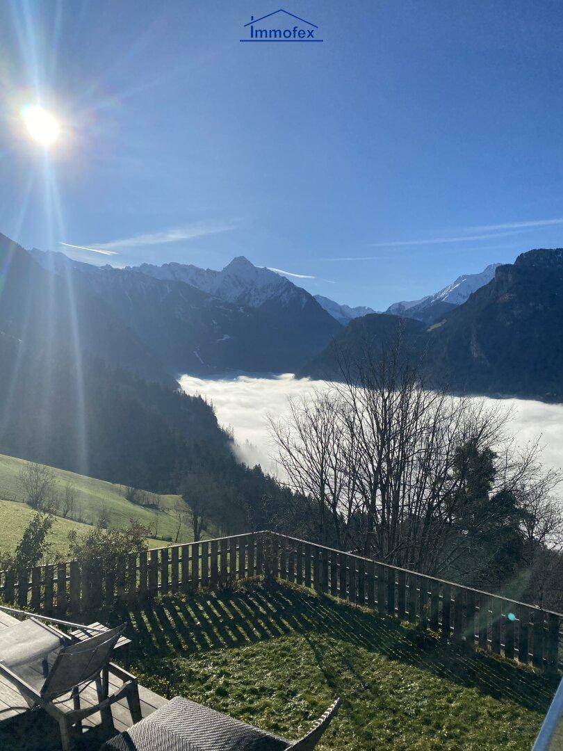 Sonniger Gartenbereich mit Holzzaun und atemberaubendem Blick auf die nebelverhangenen Berge.