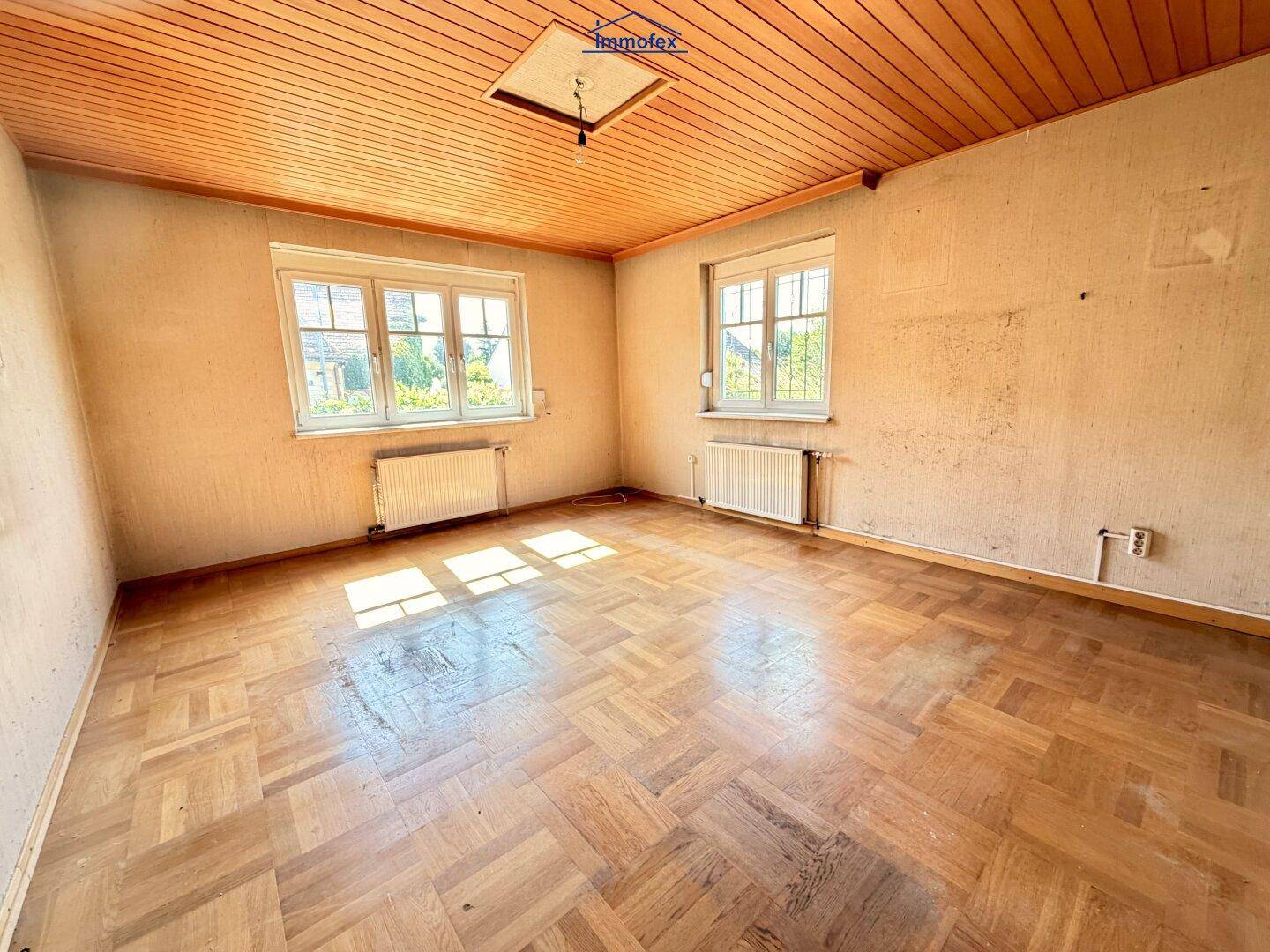 Helles Zimmer mit Fischgrätparkett, Holzdecke und zwei Fenstern, die viel Licht hereinlassen.