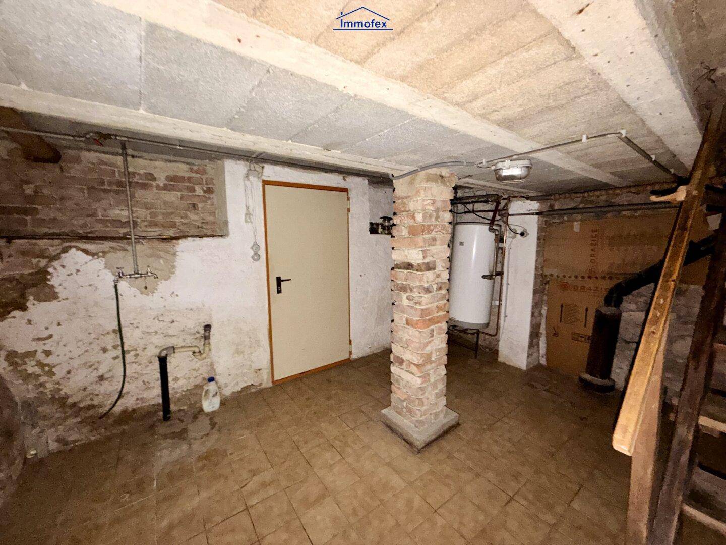 Kellerraum mit Ziegelpfeiler, Fliesenboden und Warmwasserboiler, rustikaler Zustand.