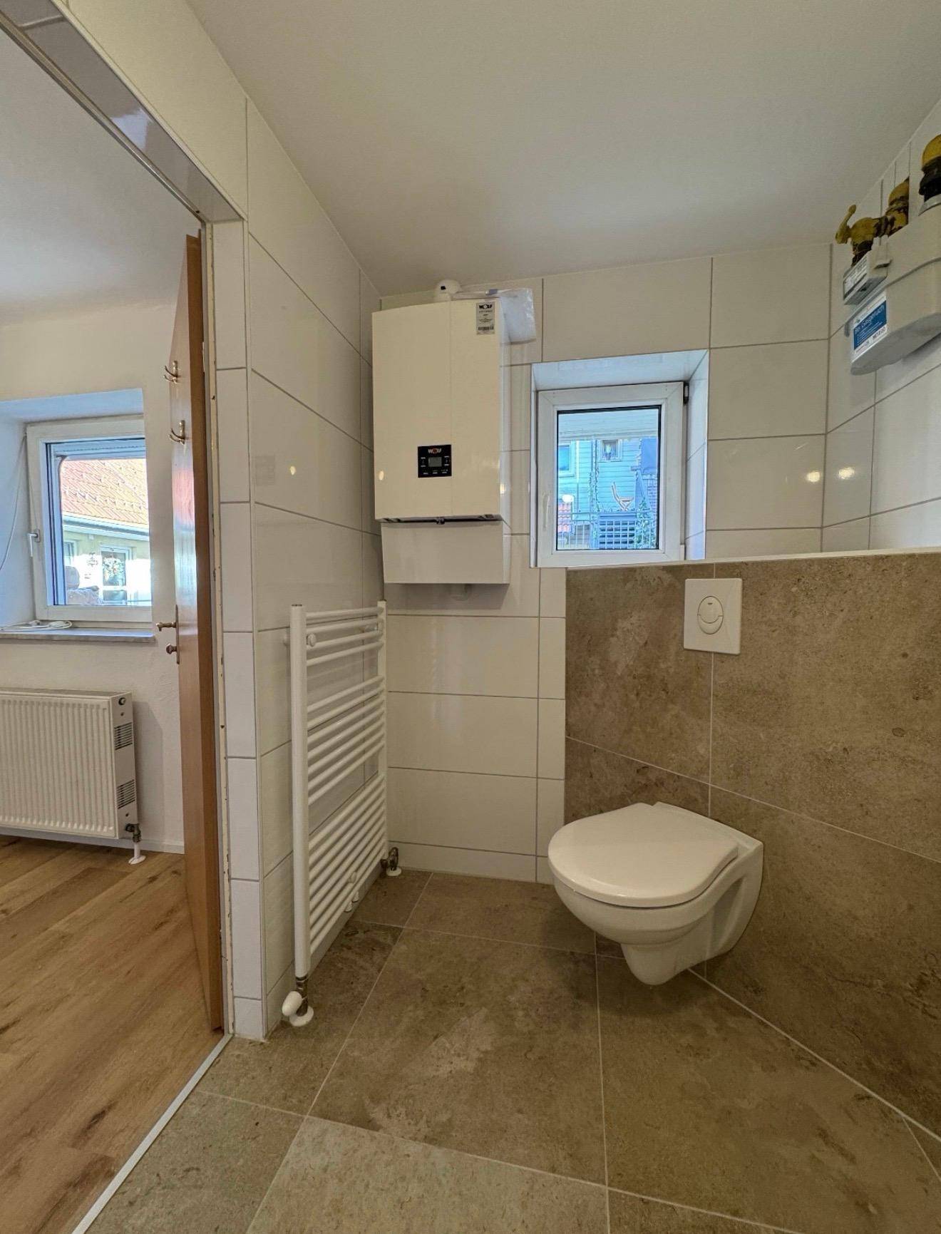 Detailansicht des Badezimmers mit wandhängendem WC, Heizkörper und Fenster für natürliche Belüftung.