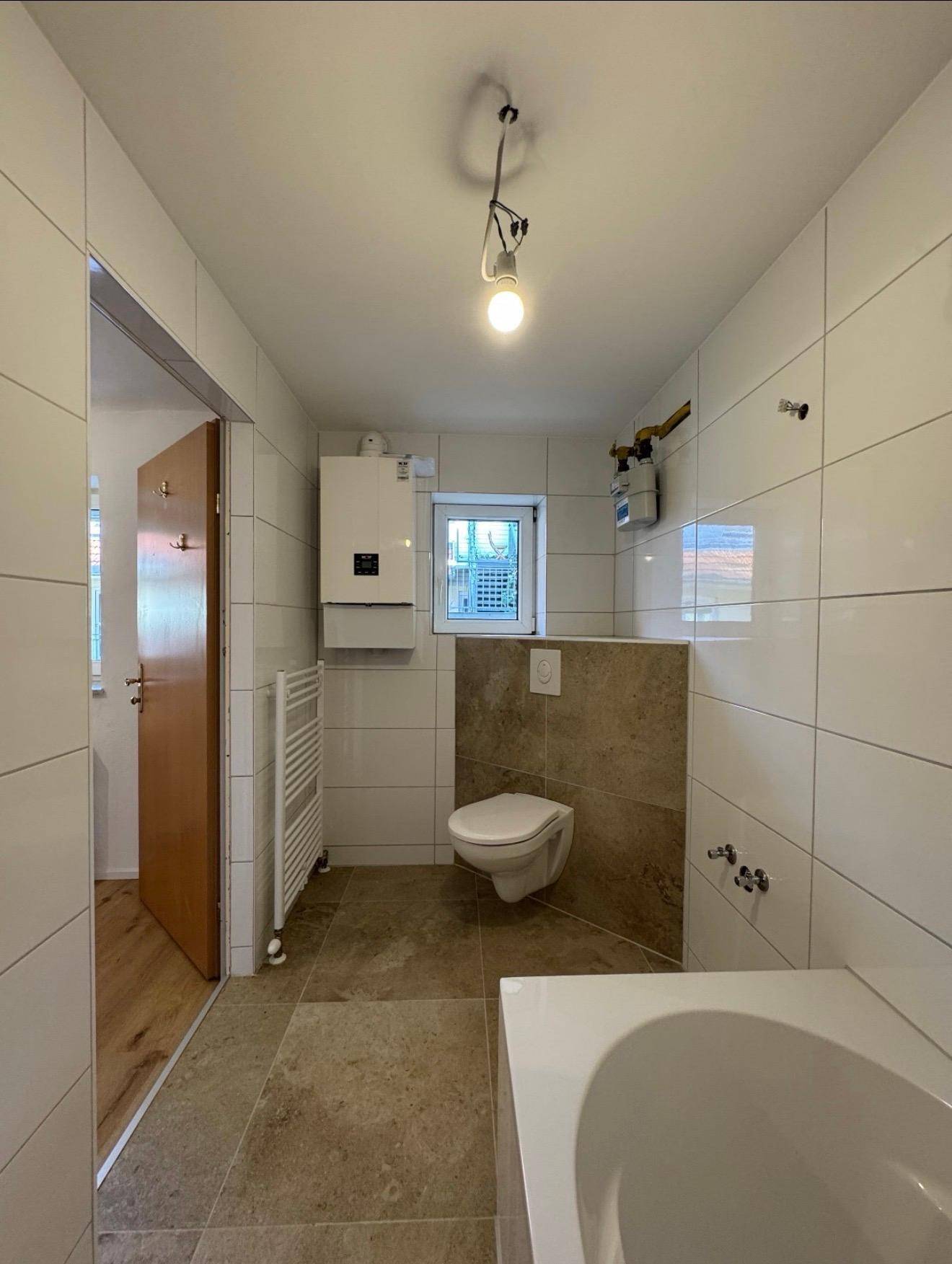 Helles Badezimmer mit WC, Fenster, Heizkörper und Wandfliesen in Weiß und Beige.
