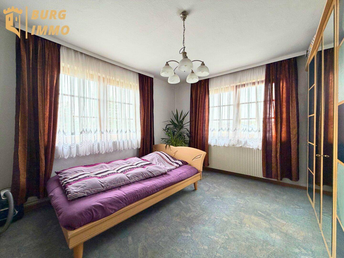 Schlafzimmer mit Teppichboden, großem Bett und zwei Fenstern mit Vorhängen.