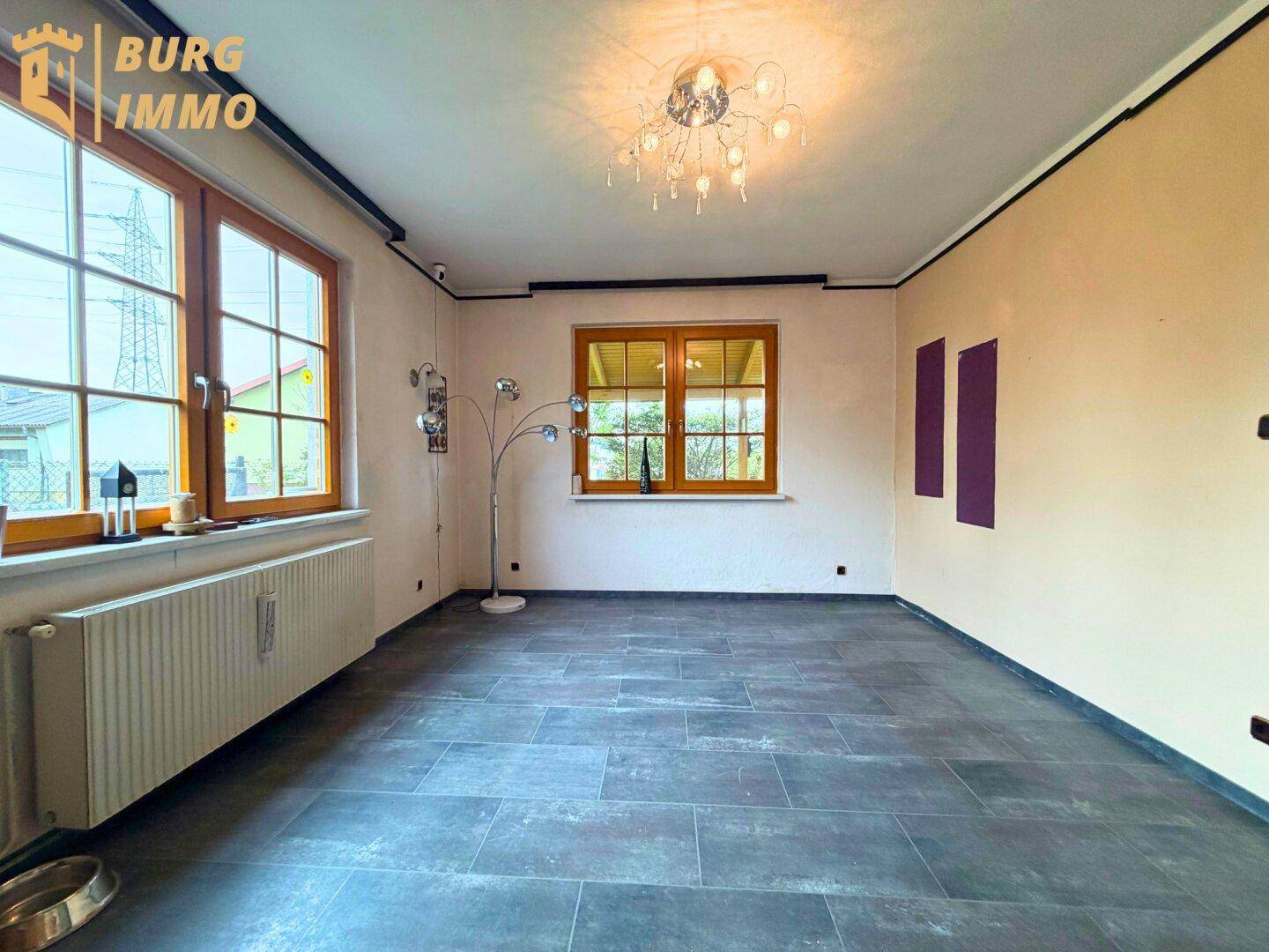 Leerer Raum mit Fliesenboden, großen Fenstern und Heizkörpern.