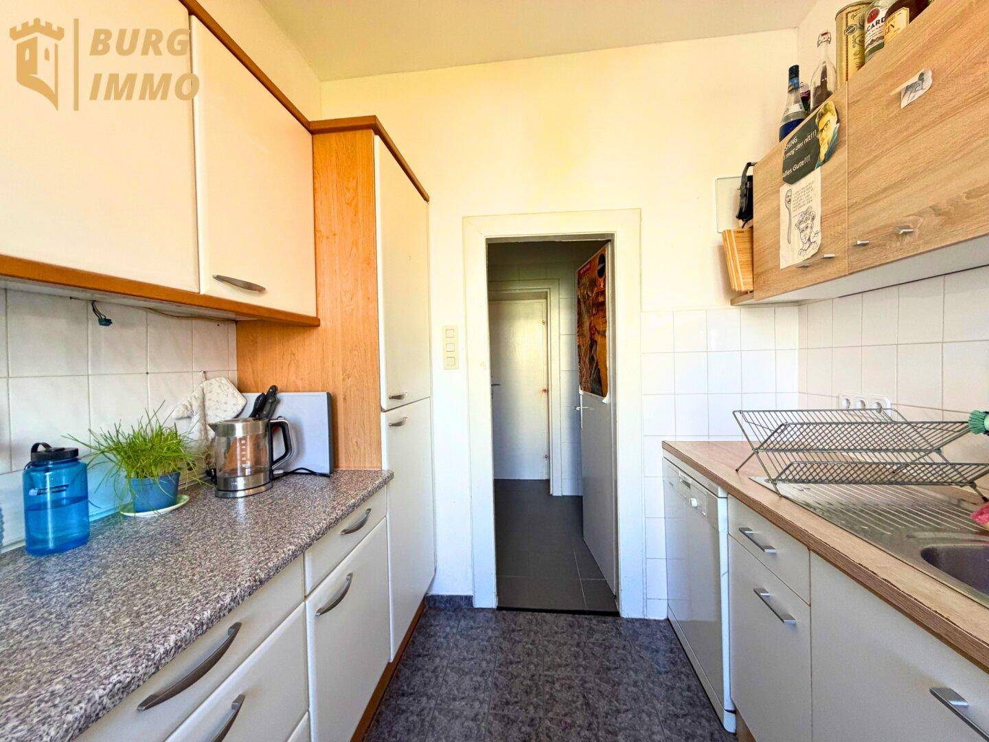 Schmale Küche mit weißen Schränken, Arbeitsplatten und Blick in den Flur.