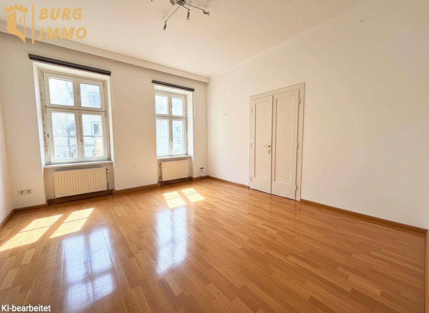 Helles Zimmer mit klassischem Parkettboden, hohen Decken und zwei großen Fenstern.