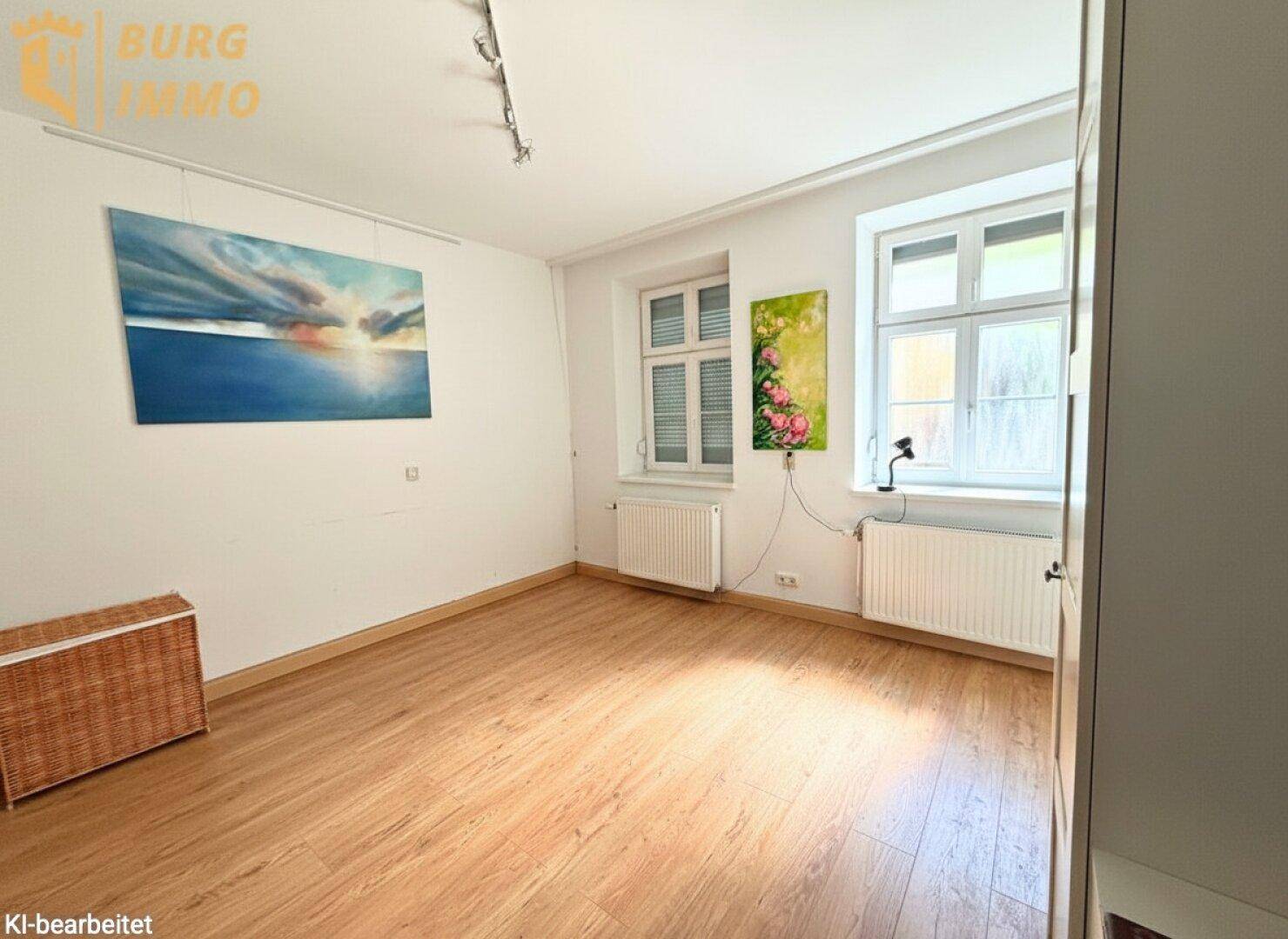 Helles Zimmer mit zwei Fenstern, Parkettboden und weiß gestrichenen Wänden.