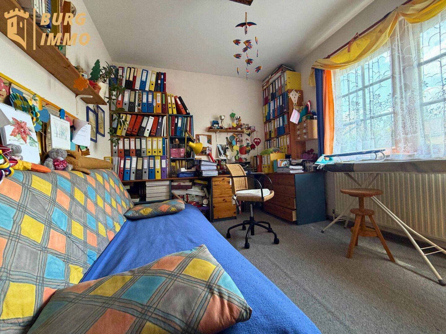 Helles Arbeitszimmer mit Teppichboden, vielen Regalen und einem großen Fenster.