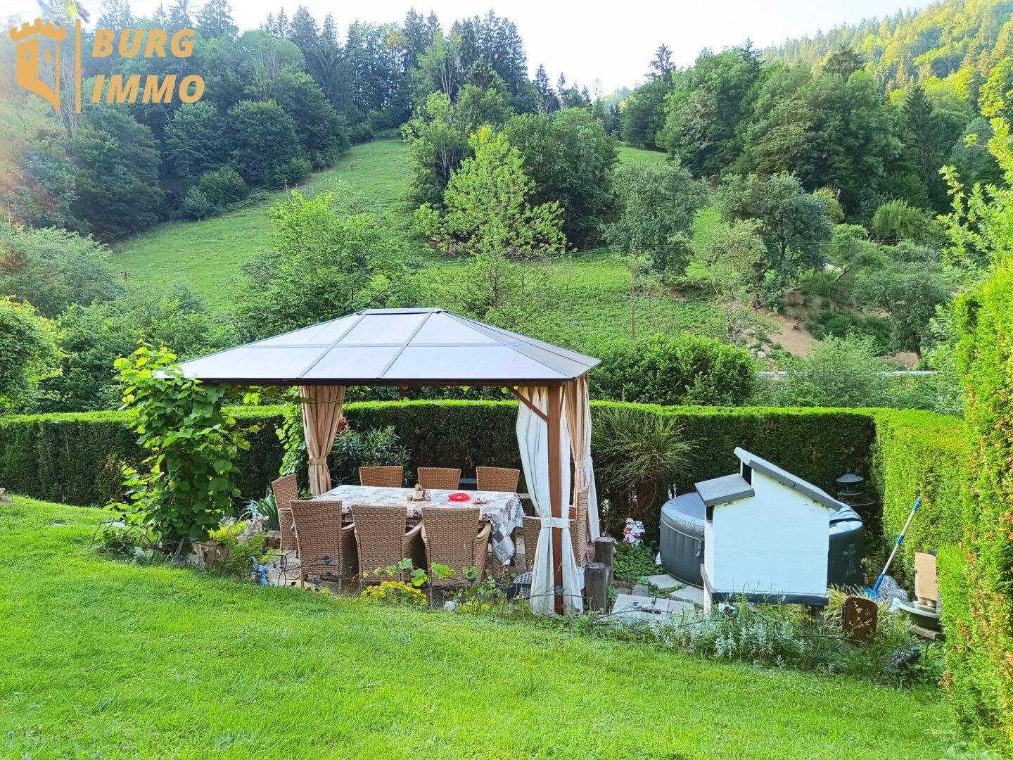 Weitläufiger Garten mit Pavillon, Essbereich und gepflegten Hecken vor grüner Hügellandschaft.