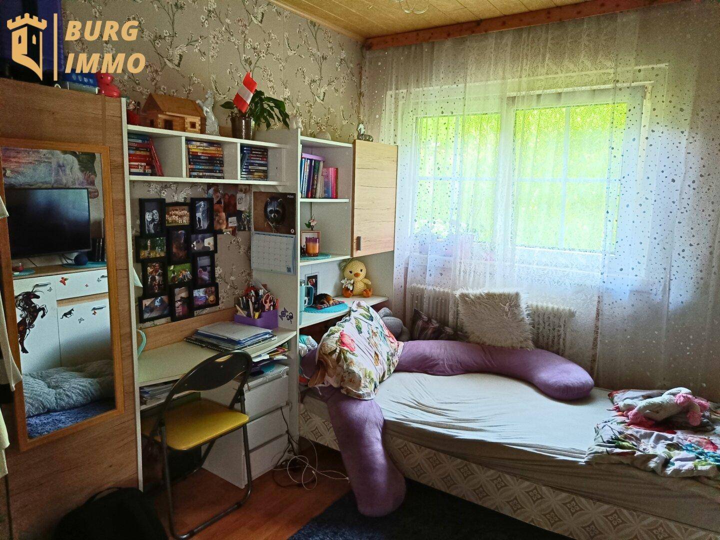 Kinderzimmer mit Einzelbett, Schreibtisch, Bücherregal und Fenster mit floraler Tapete.