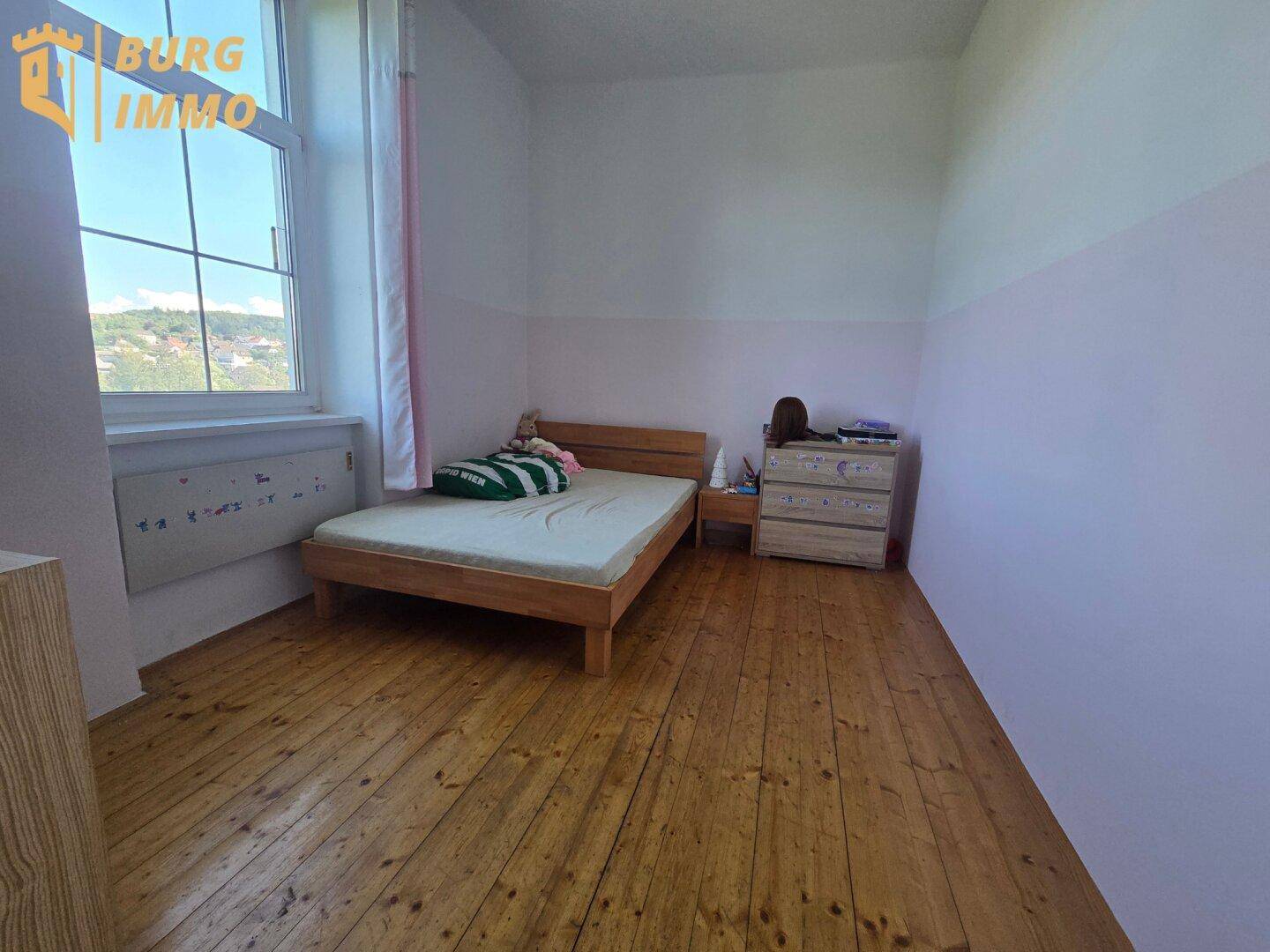 Schlafzimmer mit Holzbett, Kommode und Holzdielenboden, hell durch das große Fenster.