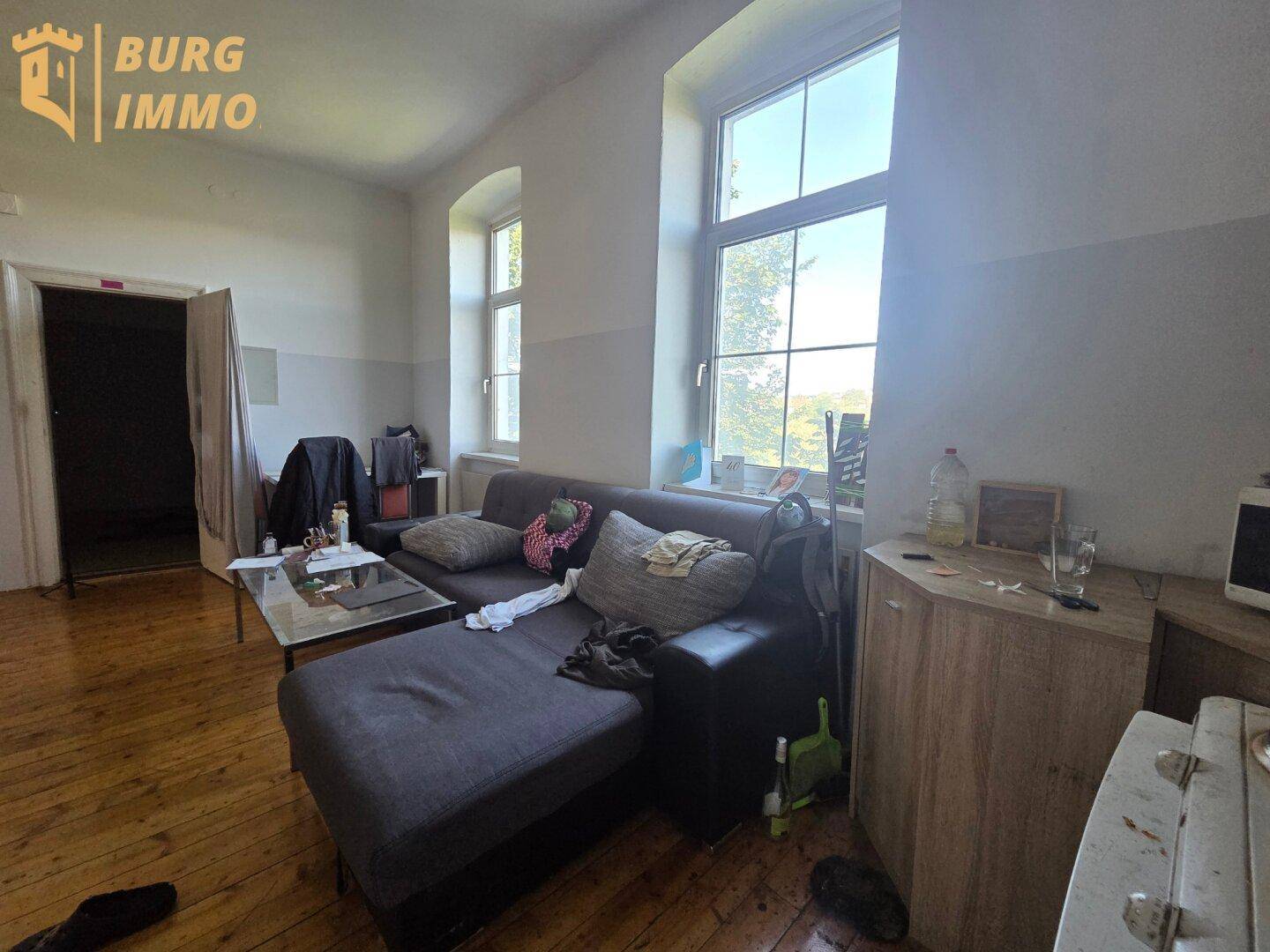Helles Wohnzimmer mit großem Fenster, Sofa und Holzdielenboden, das viel Tageslicht hereinlässt.