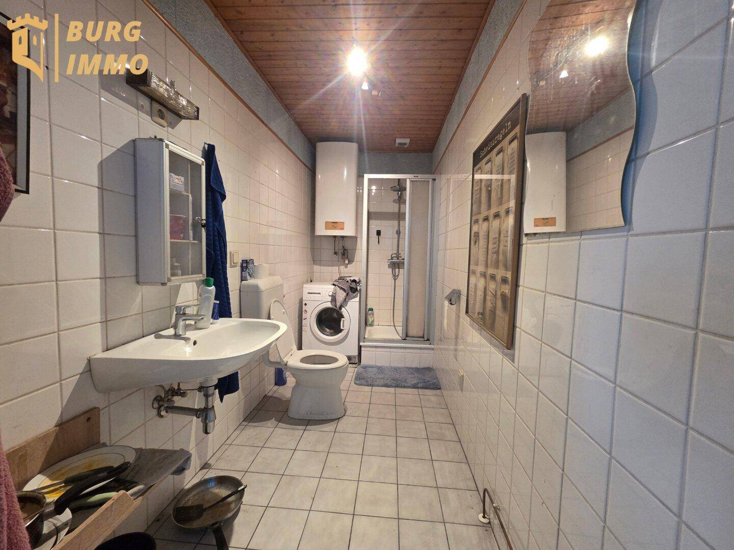 Gefliestes Badezimmer mit Waschbecken, Toilette, Dusche und Waschmaschine unter einer Holzdecke.