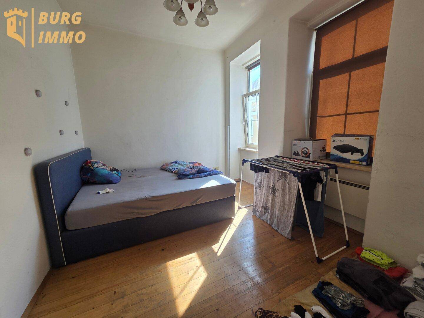 Schlafzimmer mit Einzelbett, Holzdielenboden und einem Wäscheständer, hell durch das Fenster.