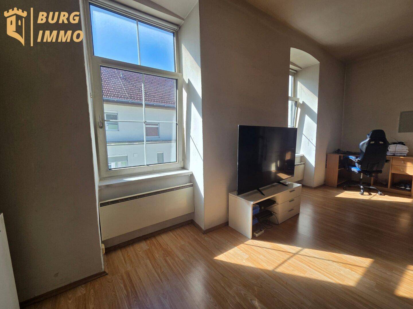 Heller Raum mit Laminatboden, großem Fenster, Fernseher und einem Arbeitsbereich.