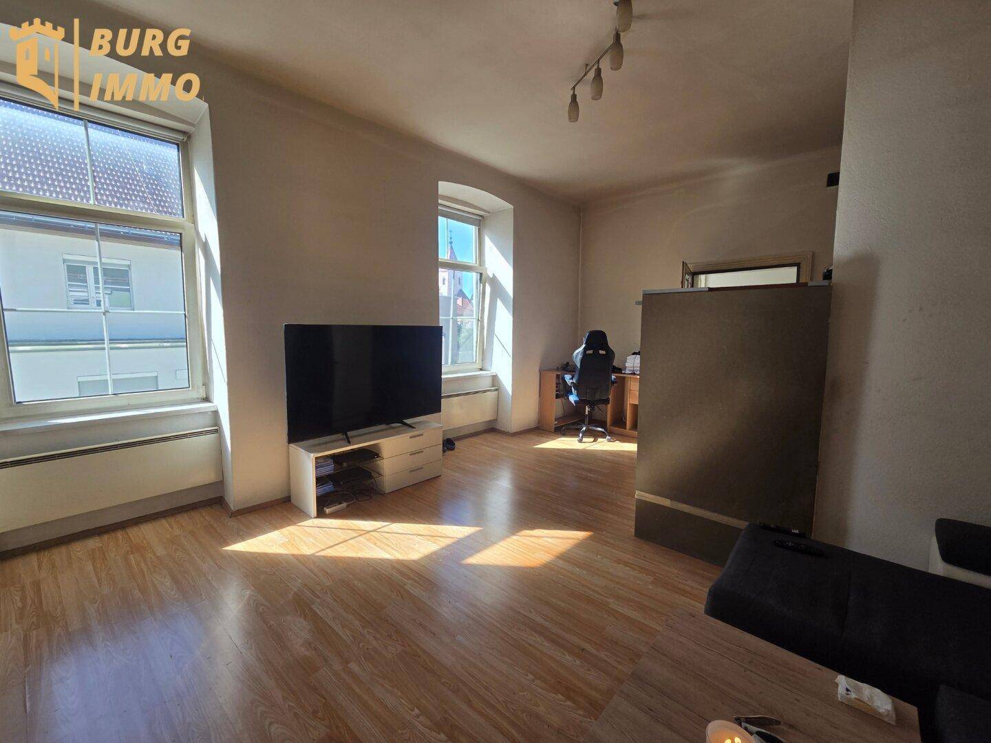 Heller Wohnraum mit Laminatboden, großem Fenster, Fernseher und einem Arbeitsbereich.