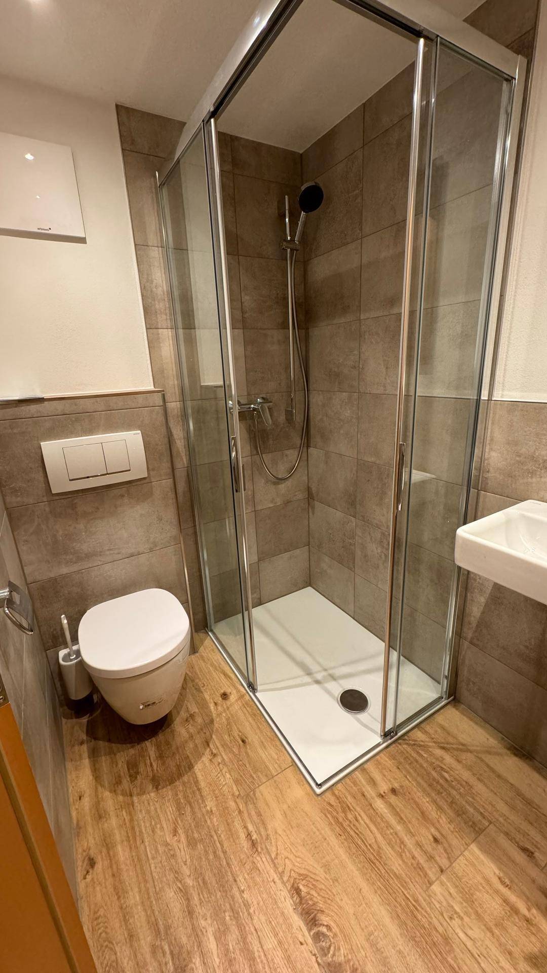 Modernes Badezimmer mit Duschkabine, WC und kleinem Waschbecken, ausgestattet mit Holzoptikfliesen.