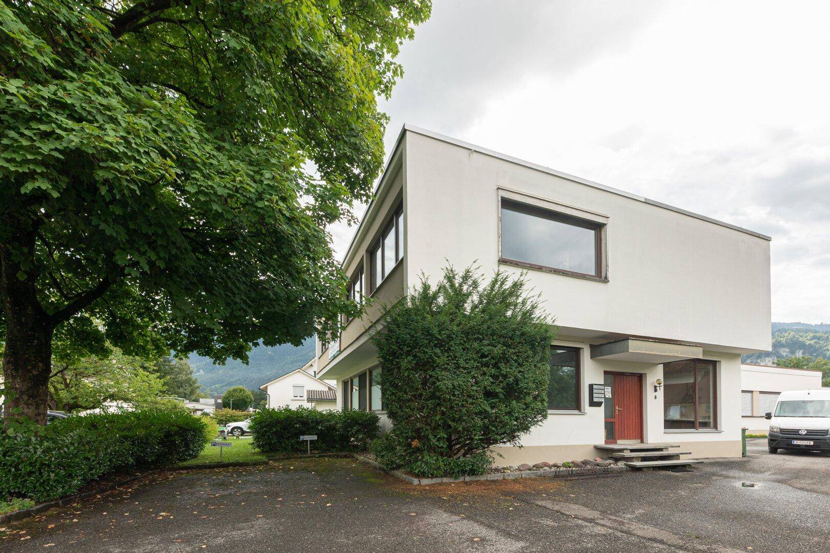 Modernes Bürogebäude mit weißer Fassade, umgeben von Bäumen und einem Parkplatz.