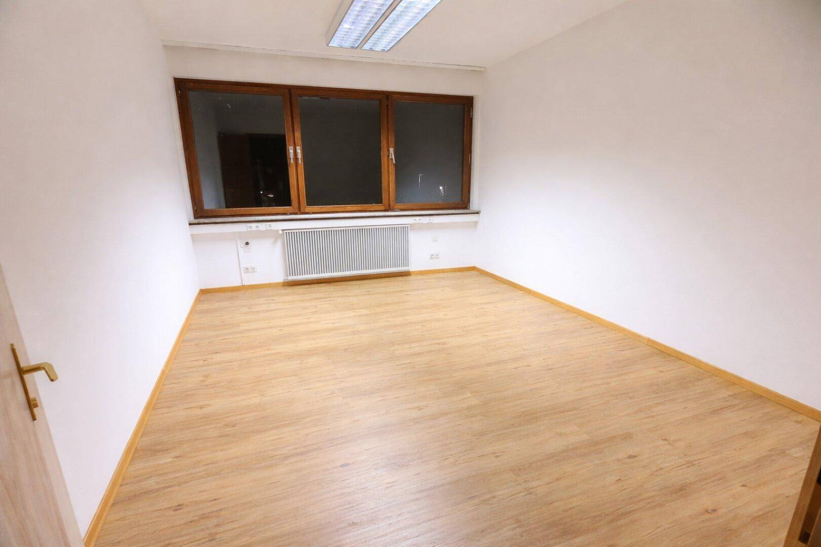 Leerer Raum mit hellem Laminatboden, weißen Wänden und großem Fenster mit Holzrahmen.