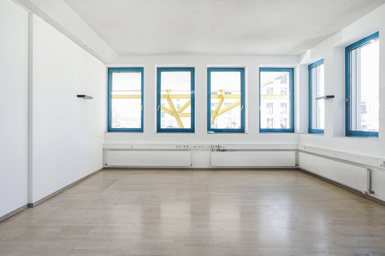 Heller Büroraum mit mehreren Fenstern, die viel Tageslicht hereinlassen, und einem gepflegten Holzfußboden.