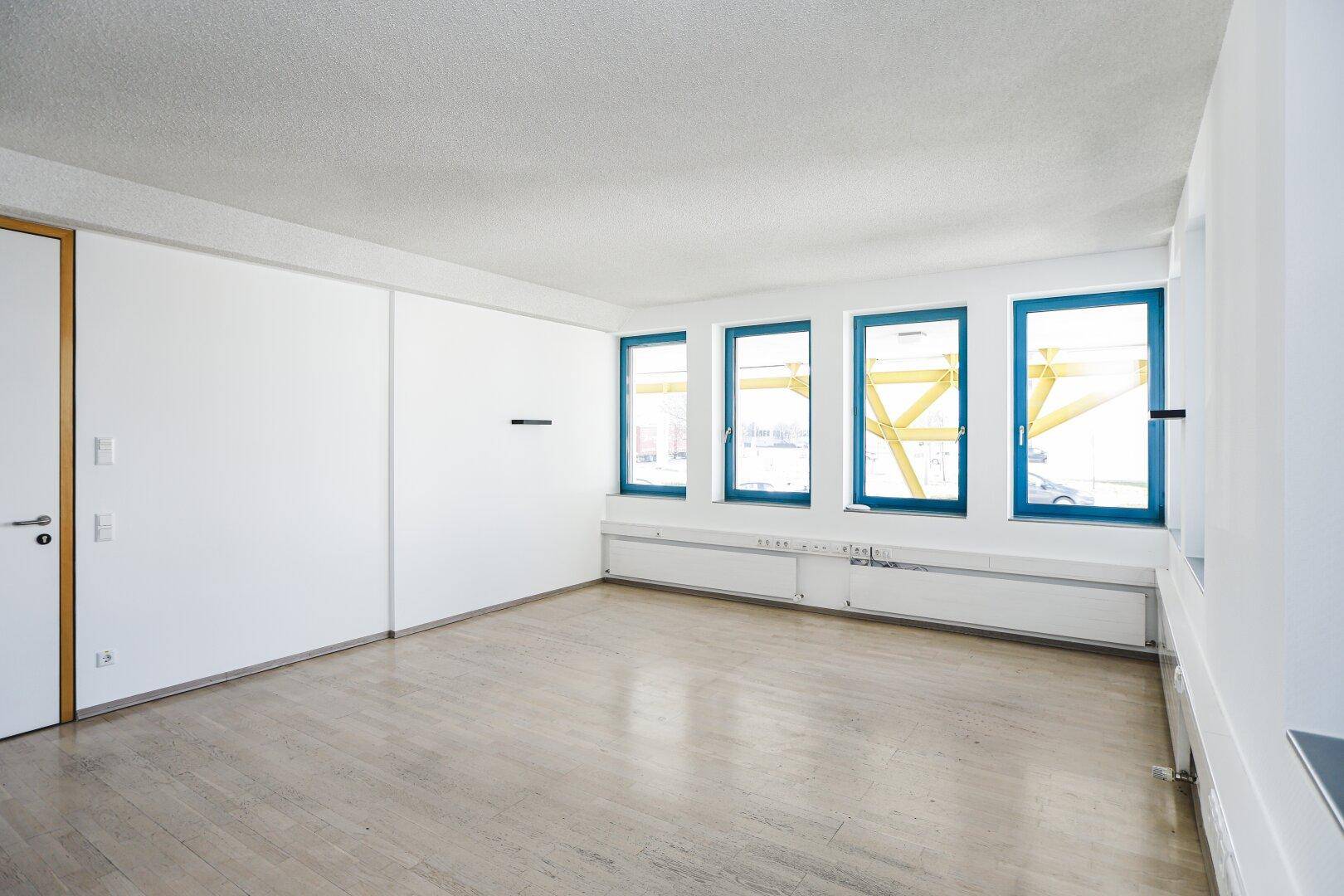 Geräumiger Büroraum mit weiß gestrichenen Wänden, hellen Böden und einer Reihe von Fenstern.