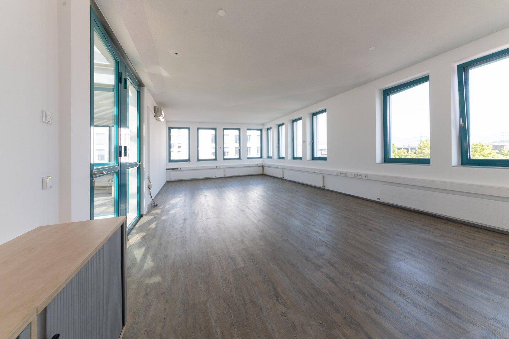 Geräumiger Büroraum mit durchgehender Fensterfront, Laminatboden und Klimaanlage.