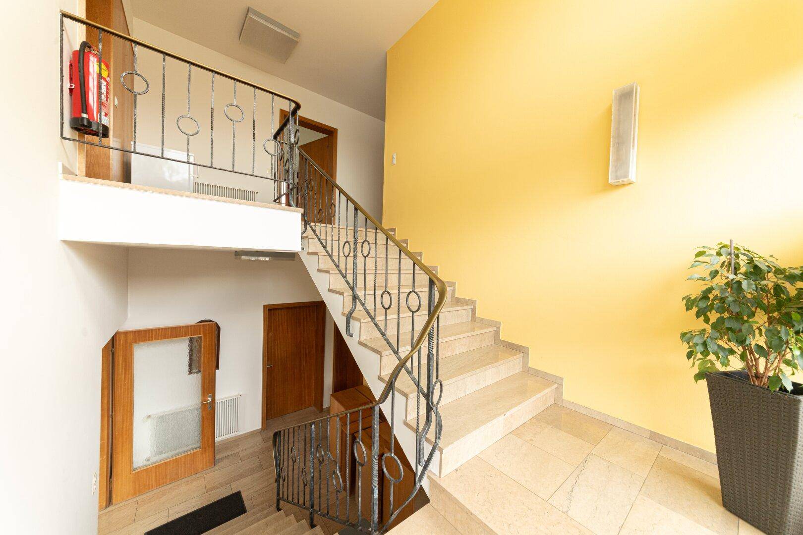 Helles Treppenhaus mit Steinfliesen, schmiedeeisernem Geländer und gelber Wand.