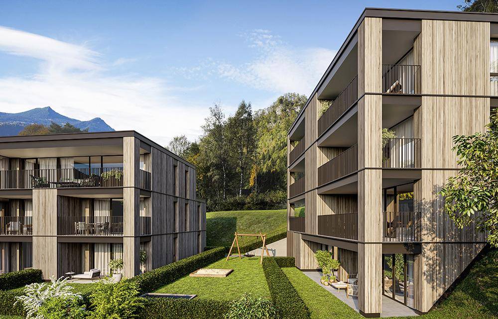 Moderne Wohngebäude mit Holzfassade und Balkonen, eingebettet in eine grüne Landschaft mit Spielplatz und Bergblick.