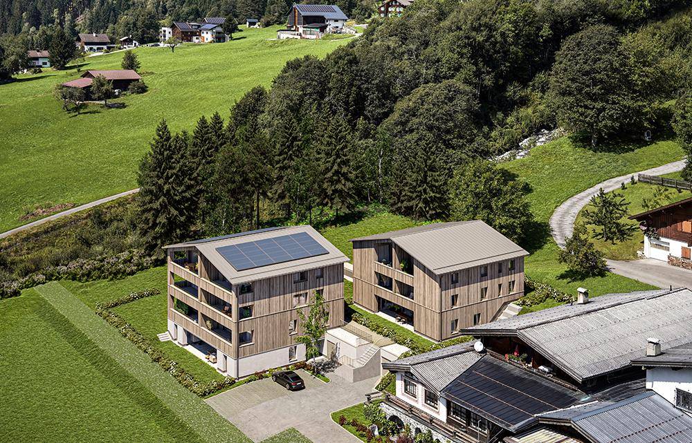 Luftaufnahme von zwei modernen Gebäuden mit Holzfassaden und Photovoltaikanlagen, umgeben von grüner Landschaft.