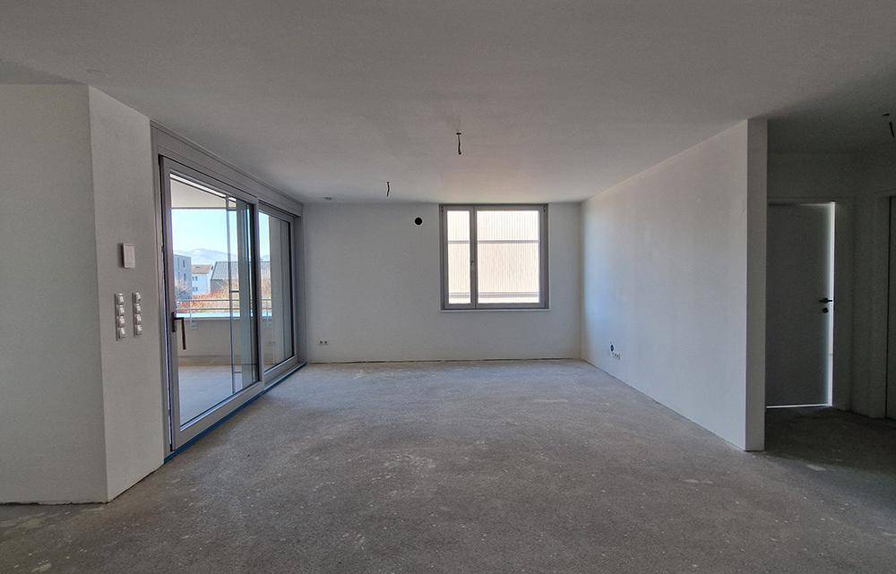 Großer, unmöblierter Raum im Rohbau mit Schiebetür zum Außenbereich und einem weiteren Fenster.