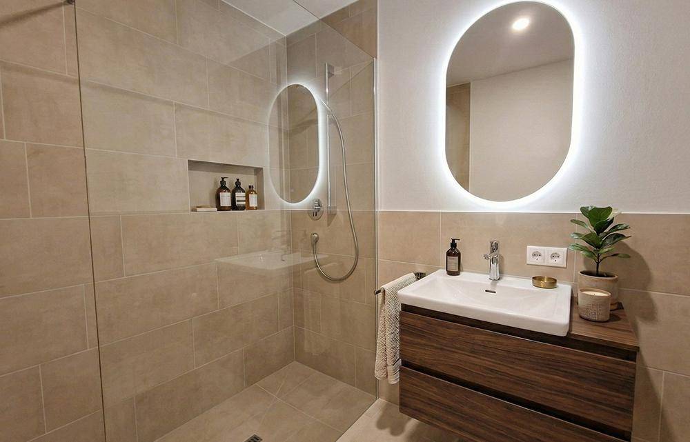 Modernes Badezimmer mit begehbarer Dusche, beleuchtetem Spiegel und beigen Fliesen.
