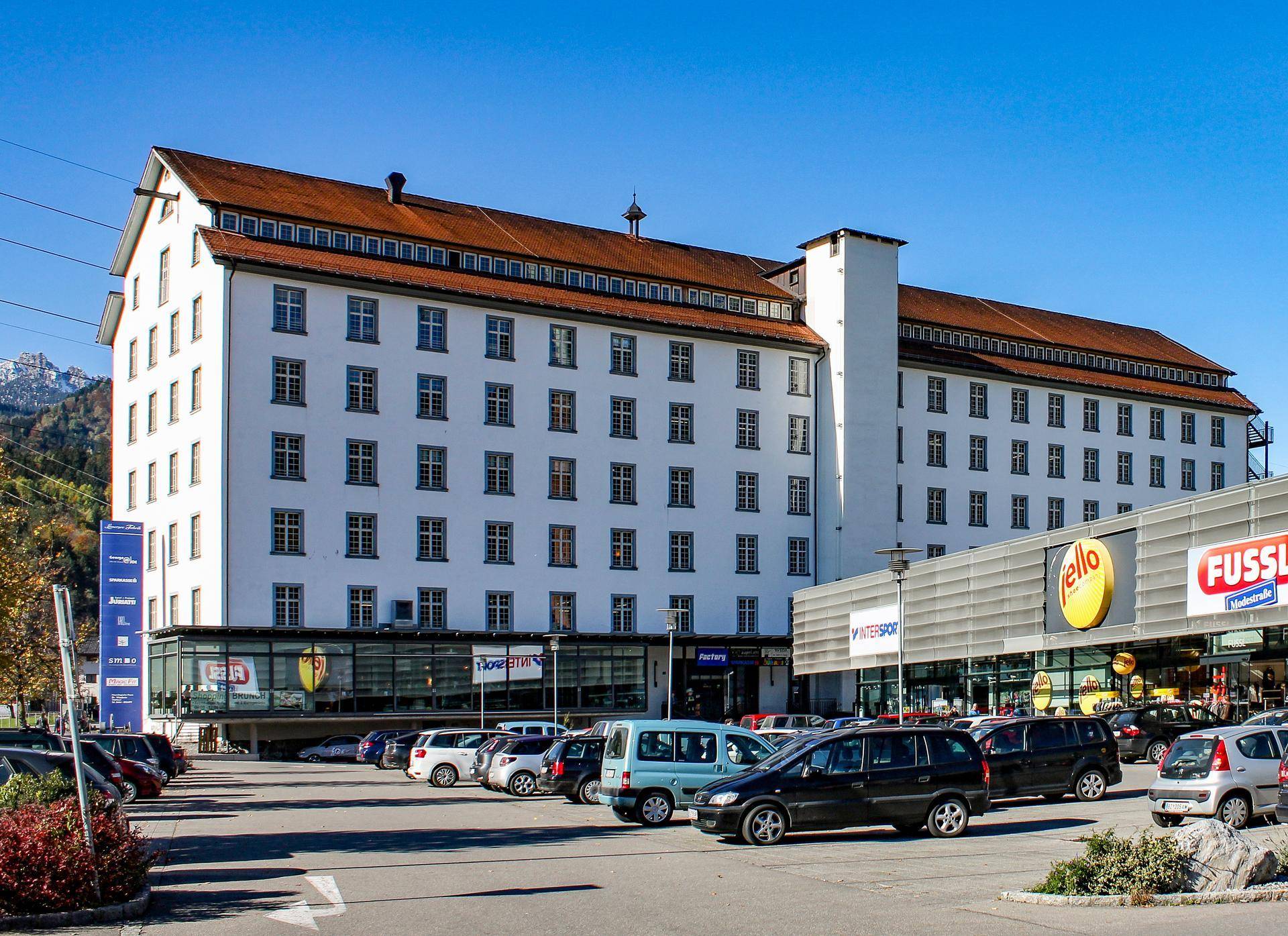 Großes, weißes Gebäude mit vielen Fenstern und einem Parkplatz davor, umgeben von Bergen unter blauem Himmel.