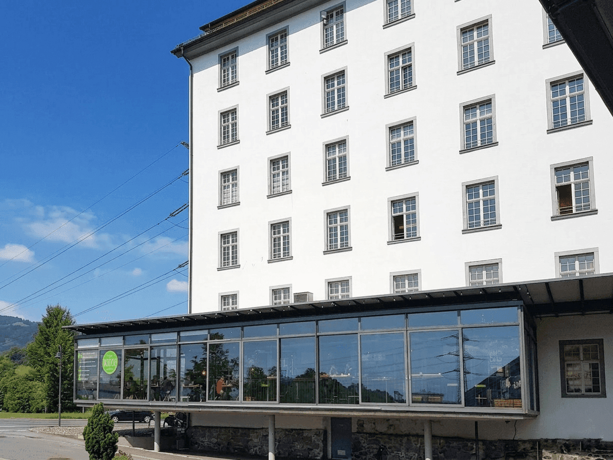 Detailansicht der weißen Fassade eines Gebäudes mit einer modernen Glaserweiterung im Erdgeschoss und grüner Umgebung.