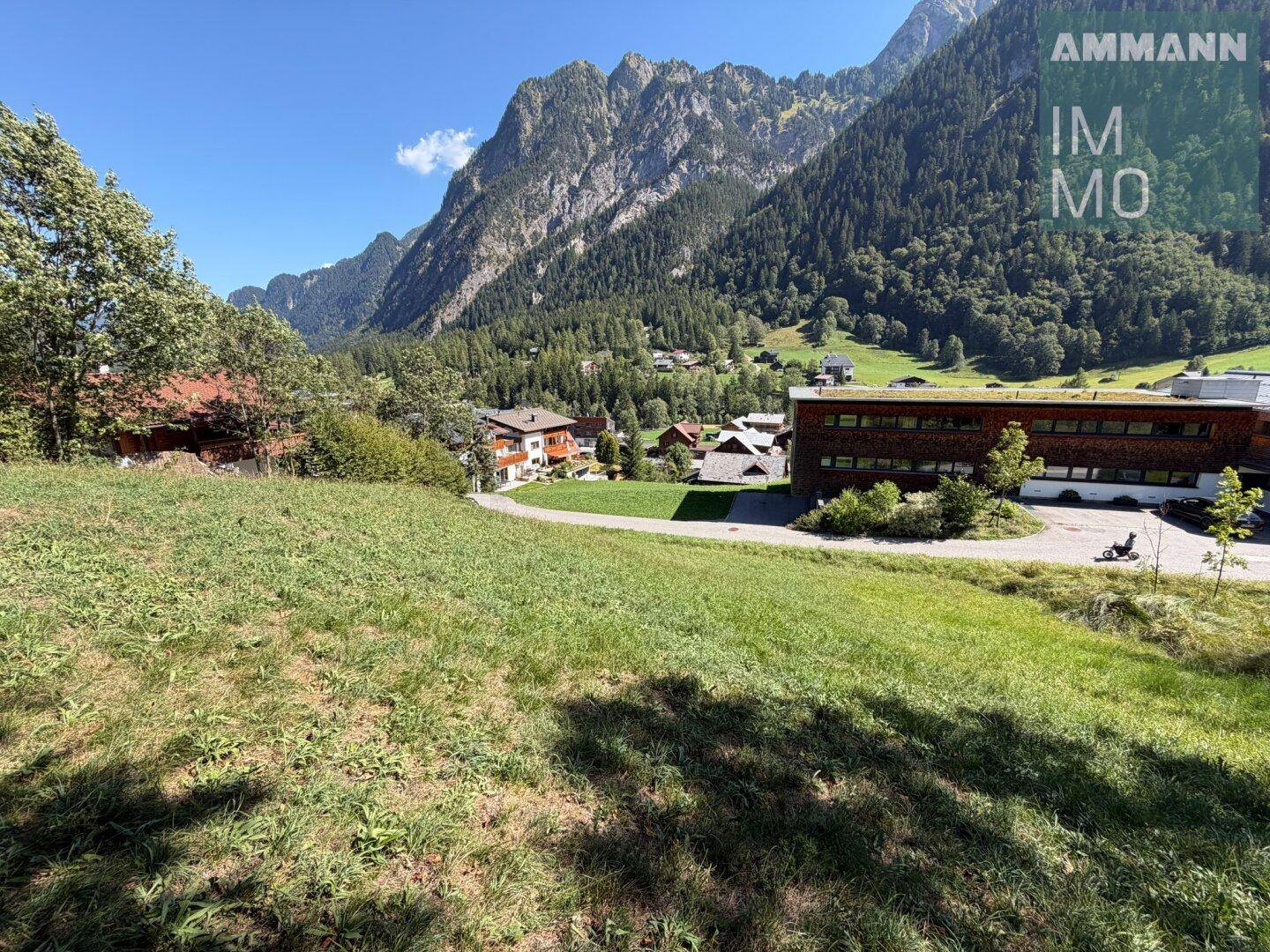 Weitläufiger Blick über grüne Wiesen und das Tal mit traditionellen Häusern vor imposanter Bergkulisse.