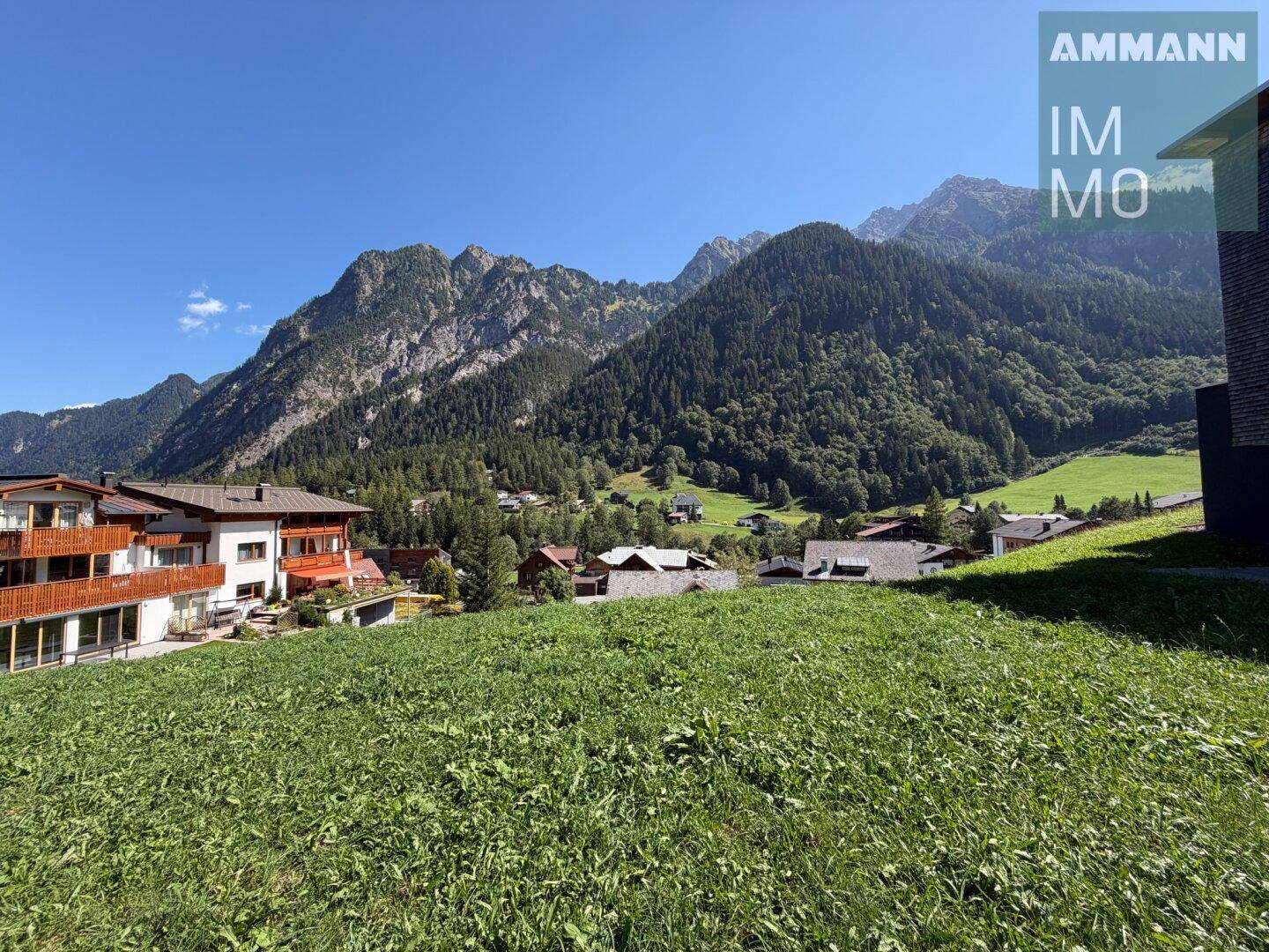 Blick von einer grünen Anhöhe auf moderne Gebäude und die majestätische Bergwelt im Hintergrund.