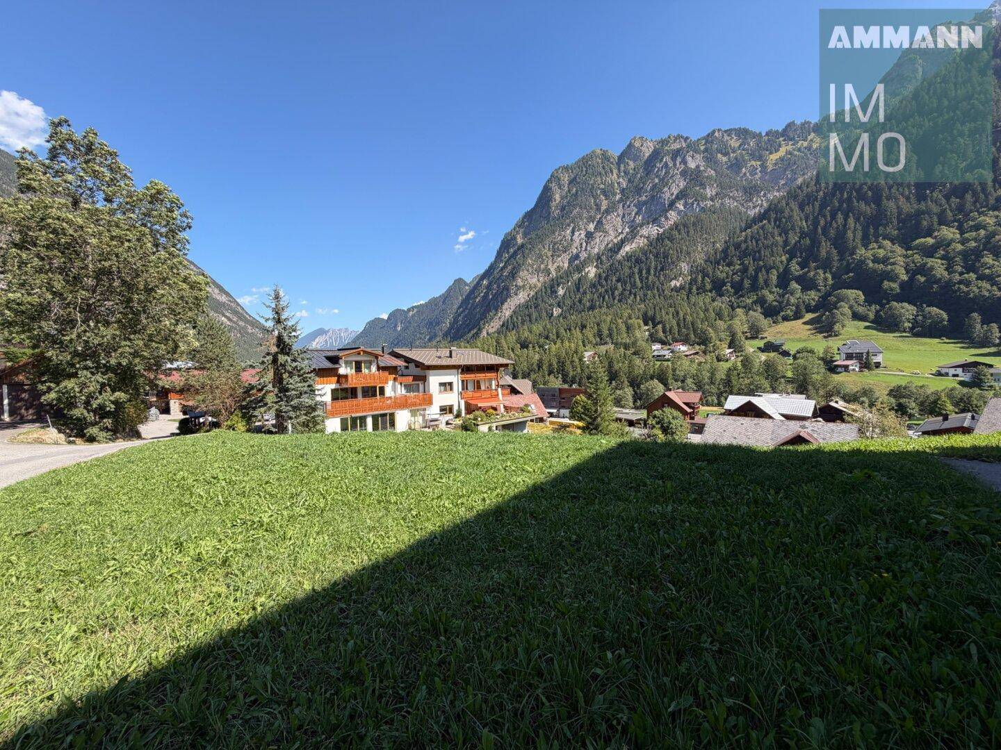 Weitblick über eine grüne Wiese auf ein Mehrfamilienhaus mit Balkonen und die umliegenden Berge.
