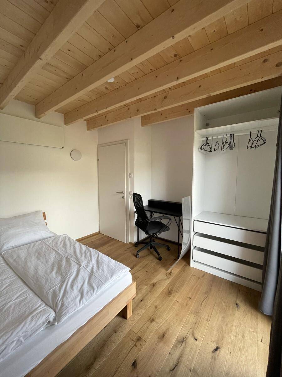 Schlafzimmer mit Holzbalkendecke, Schreibtisch und offenem Kleiderschrank für Stauraum.