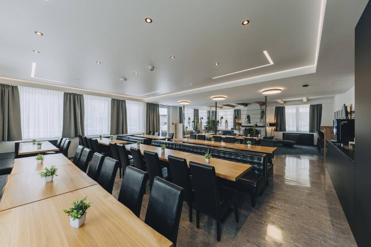 Weitläufiger Speisesaal mit zahlreichen Holztischen und schwarzen Sitzgelegenheiten, der in einen gemütlichen Loungebereich übergeht.