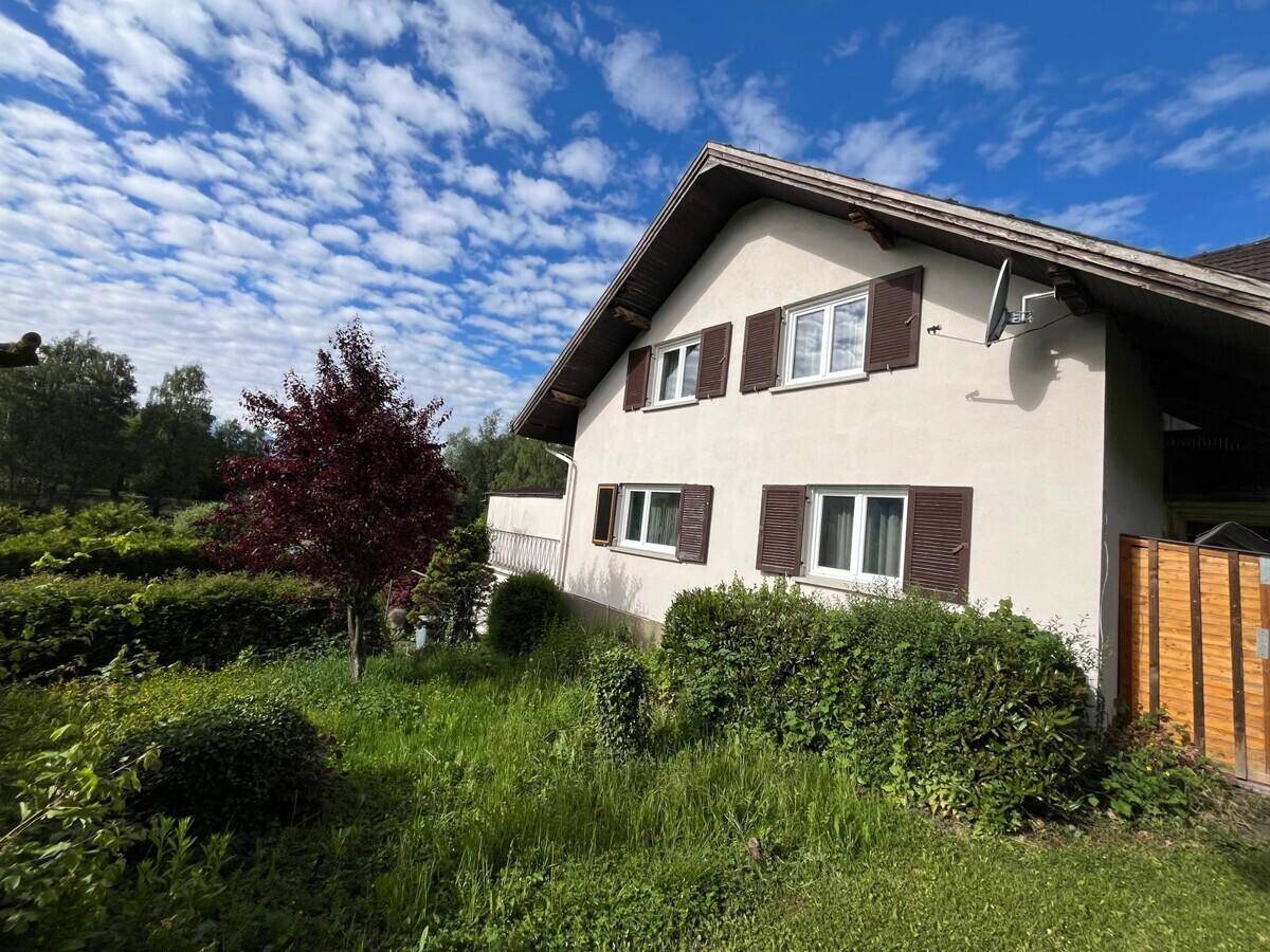 Seitenansicht des Hauses mit heller Fassade, Fenstern mit Fensterläden und üppiger Bepflanzung.