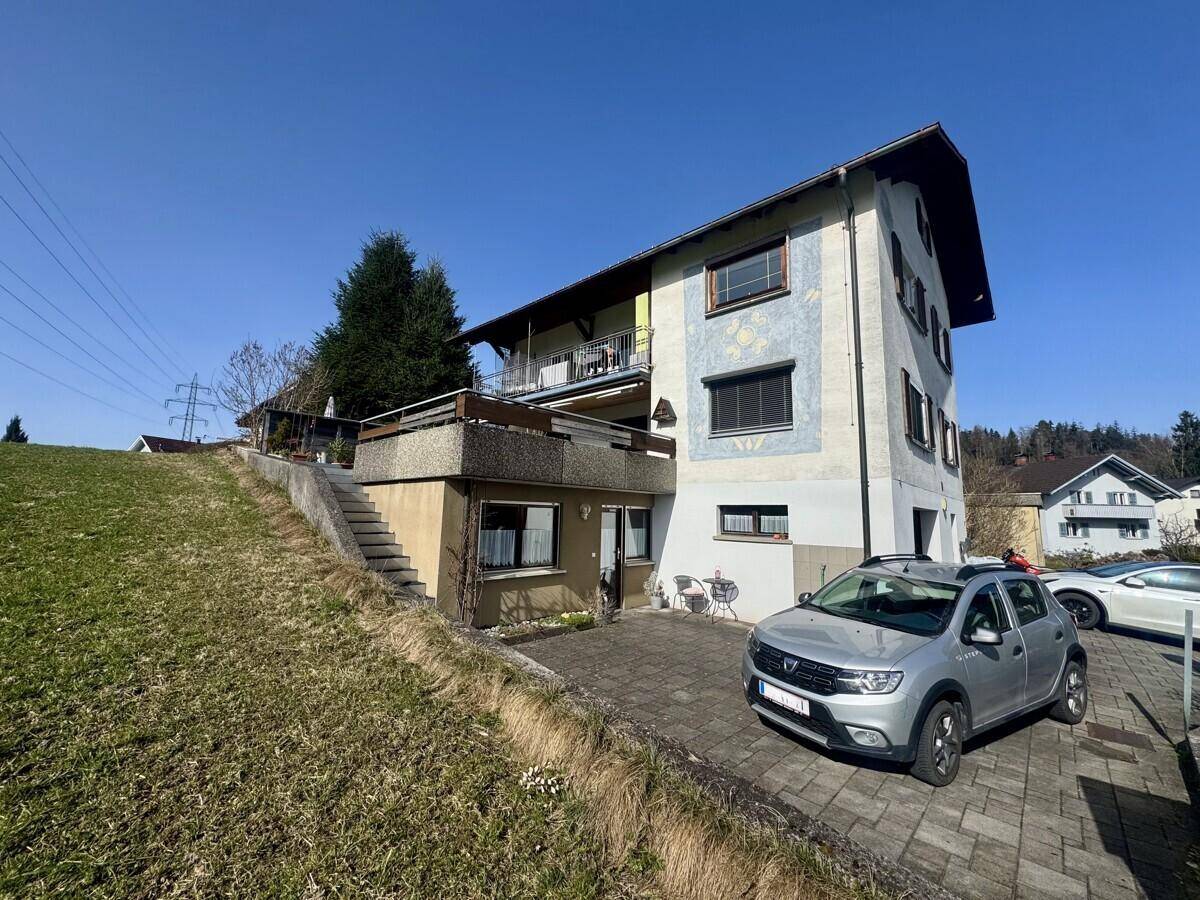 Mehrstöckiges Wohnhaus an einem Hang mit Balkonen und weitem Bergblick unter blauem Himmel.